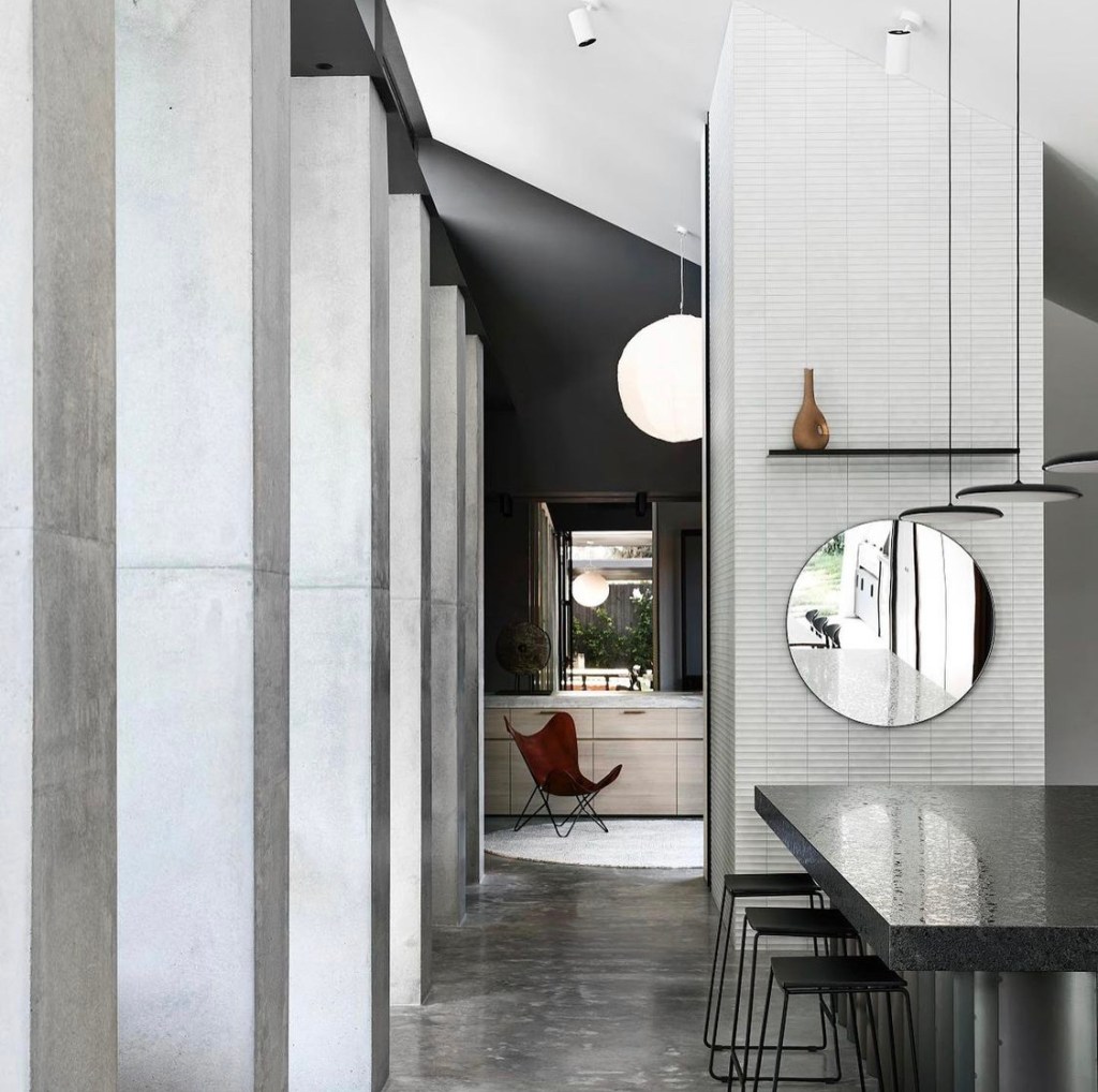 Modern interior showcasing a sleek corridor with concrete pillars, a black granite countertop, and stylish furniture, featuring a large circular mirror and a hanging pendant light.